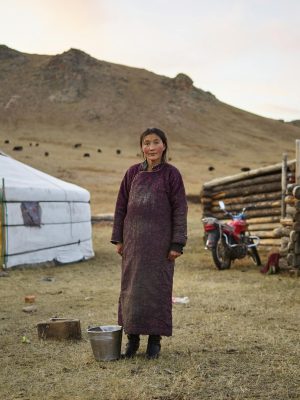 Full body of middle aged pensive Mongolian female farmer in national clothes looking at camera against yurts in valley