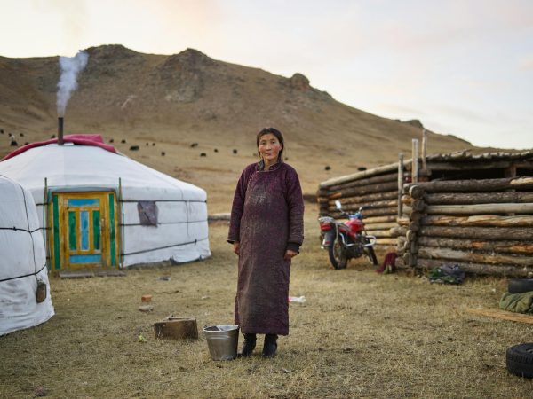 Full body of middle aged pensive Mongolian female farmer in national clothes looking at camera against yurts in valley